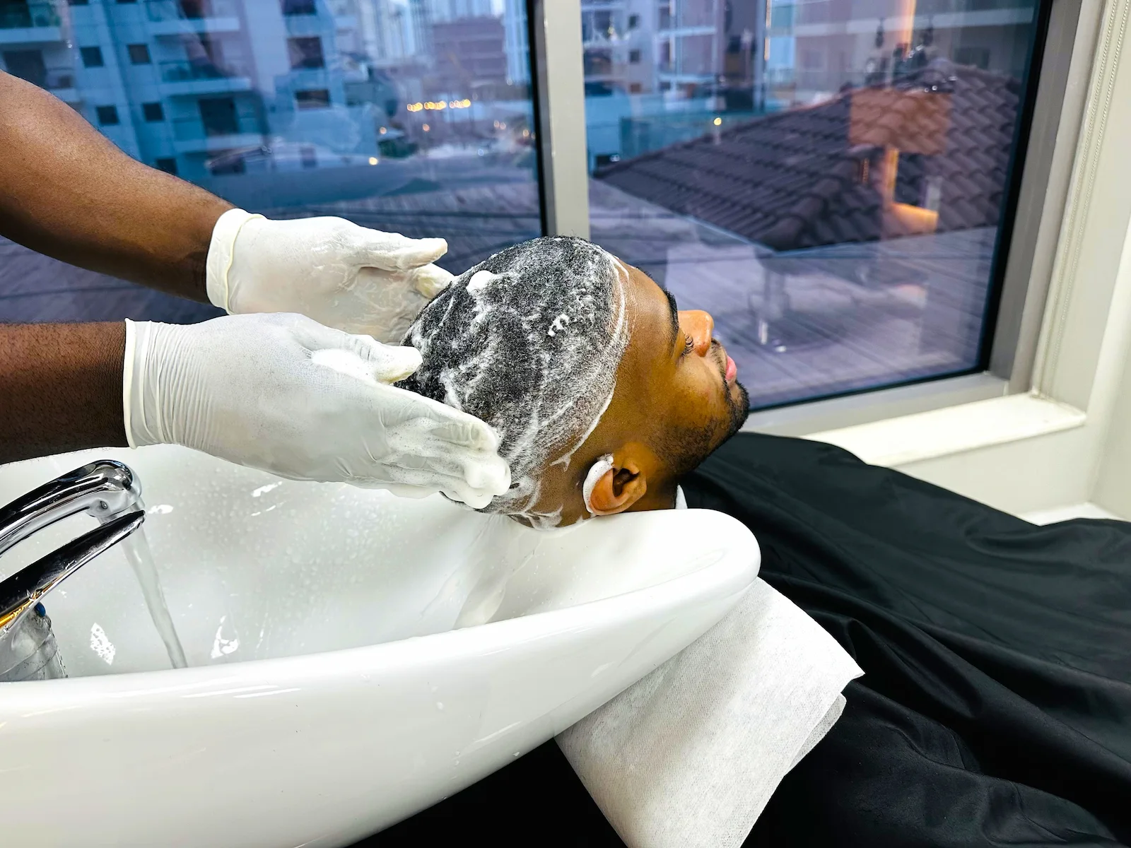 man getting his hair washed at a barbershop sink, barber wearing gloves applying shampoo and massaging scalp